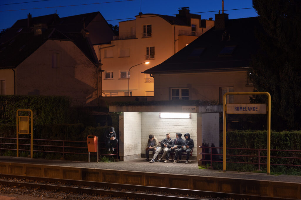 Teenagers sitting in an illuminated train stop, with Luxembourg town buildings behind them at night.