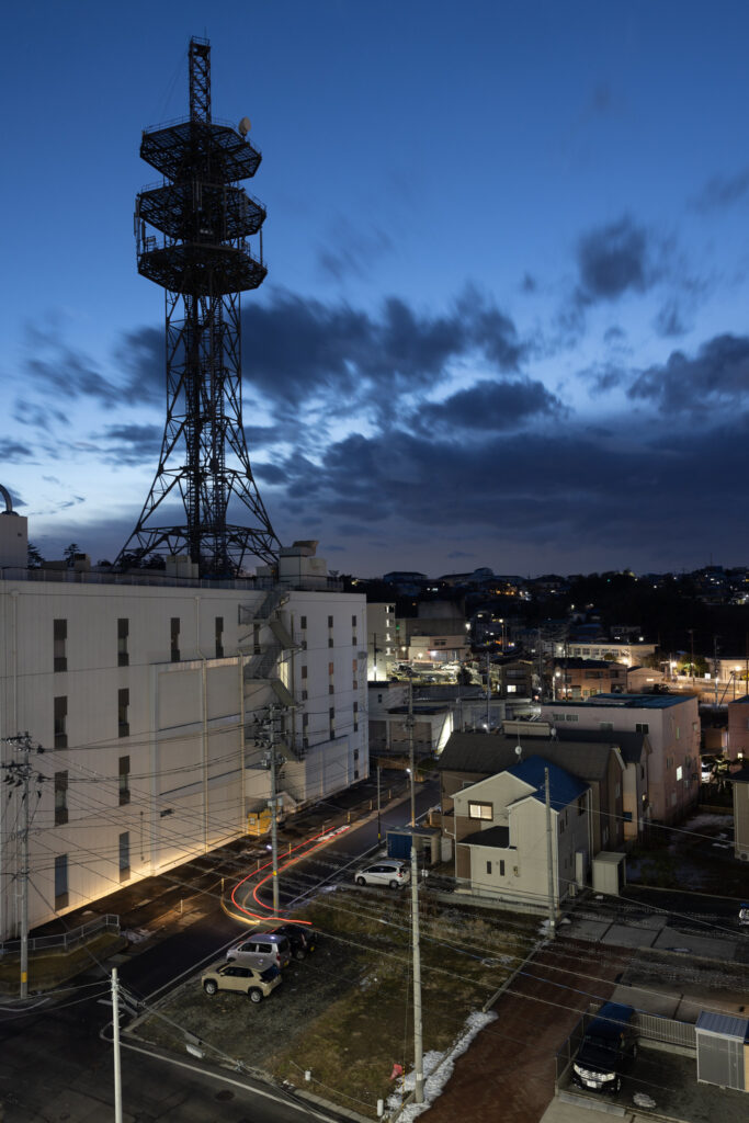 A nighttime view of a cityscape in T?hoku, Japan, featuring a tall communication tower, buildings, and streets illuminated by streetlights and vehicle lights.