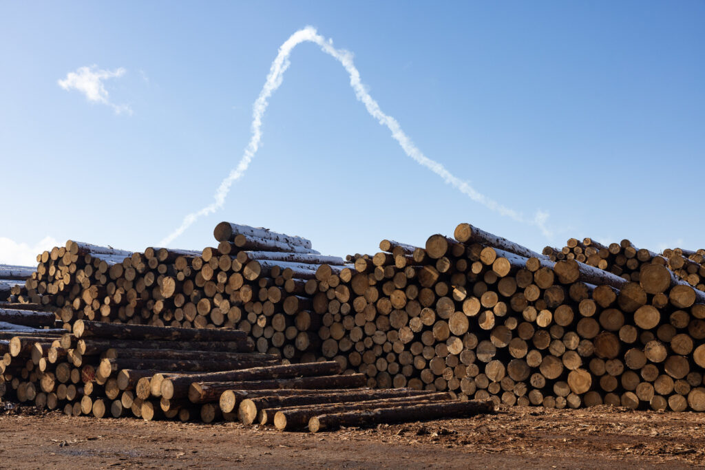 A stack of logs in a lumber yard under a clear blue sky, with white smoke trails curving in a "V" above.