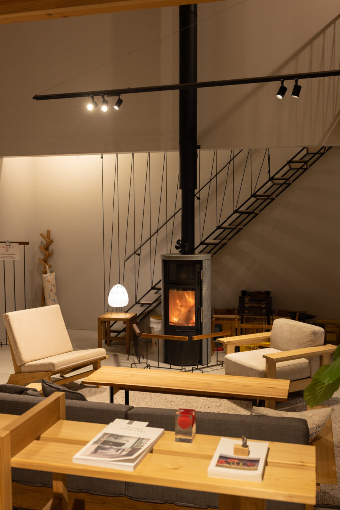 An interior space of Ishinomaki Laboratory Home Base featuring a wood-burning stove, wooden furniture, and modern lighting, with a staircase in the background.