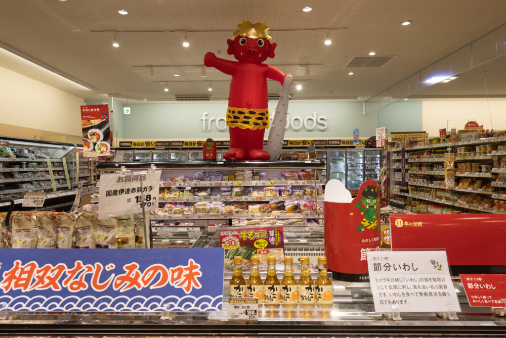 An interior view of a grocery store in Japan featuring an inflatable red demon figure on display, with various food items and signs in Japanese on shelves and counters.