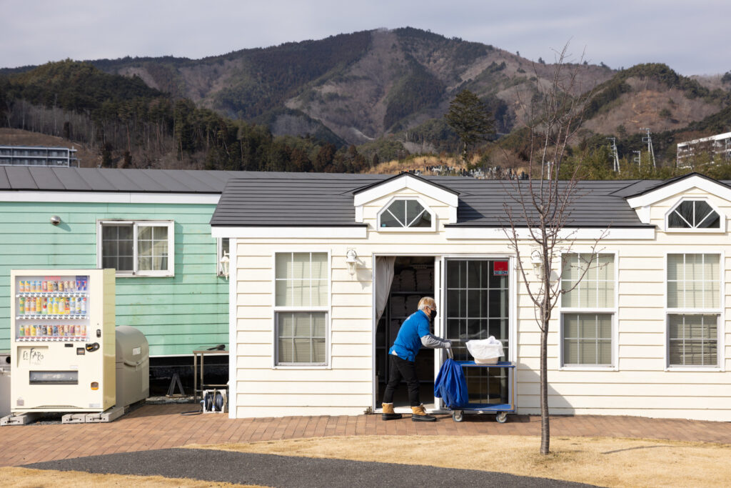 A person in a blue jacket is seen pushing a cart outside a mobile home-style hotel building. In the background, there are mountains and another building painted light green, along with a vending machine.