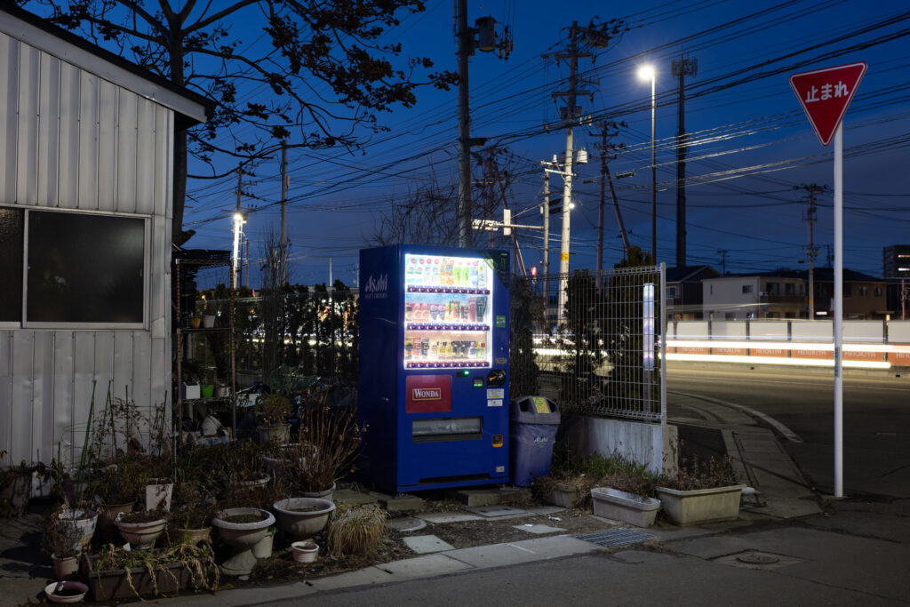 A vending machine on a corner with a yield sign and a car passing at dusk.