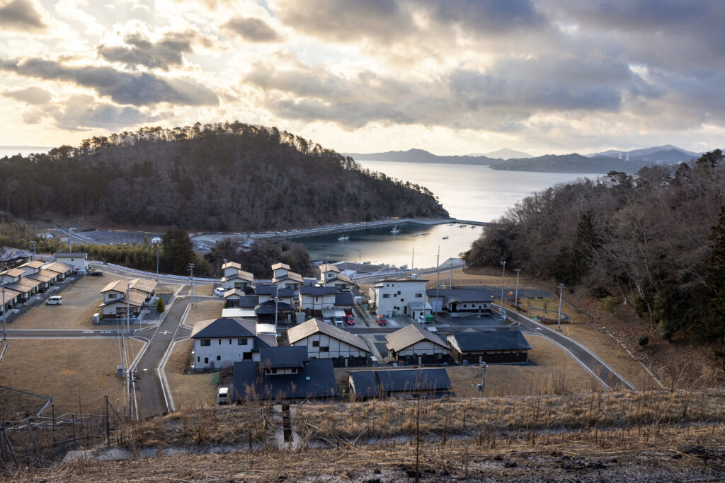 A panoramic view of a coastal settlement in T?hoku, Japan, showing a cluster of houses and roads near a harbor, with a background of hills and cloudy skies.