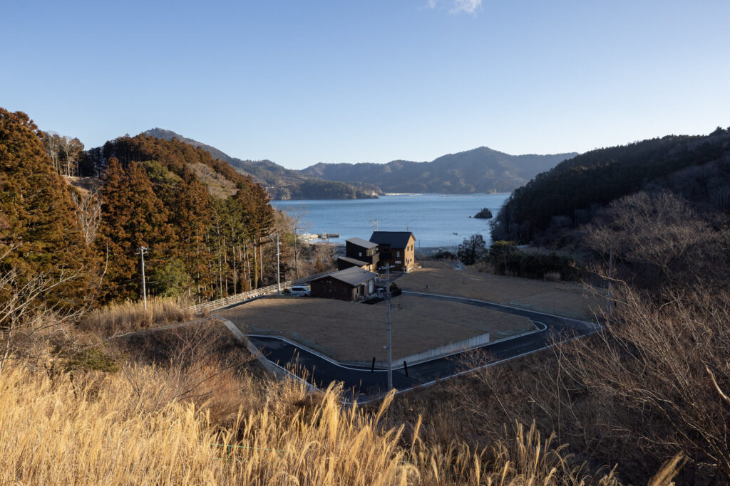 A panoramic view of a coastal landscape in T?hoku, Japan, showcasing a few buildings near the shore, surrounded by trees and a backdrop of rolling hills and mountains under a clear blue sky.