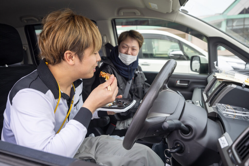 Two young men sitting inside a car, one is eating while the other is smiling and watching, with food containers visible on the dashboard.