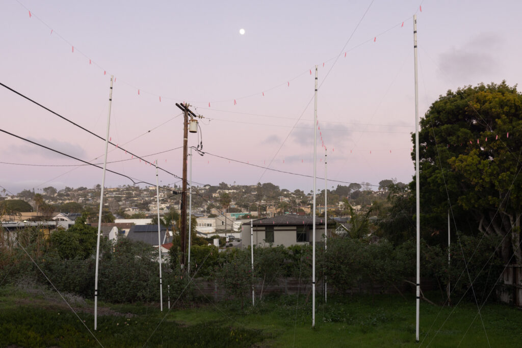 A dusk photograph of flags strung between white poles, illustrating the shape of a proposed building. The moon hangs in the sky with small houses rising up a hill.