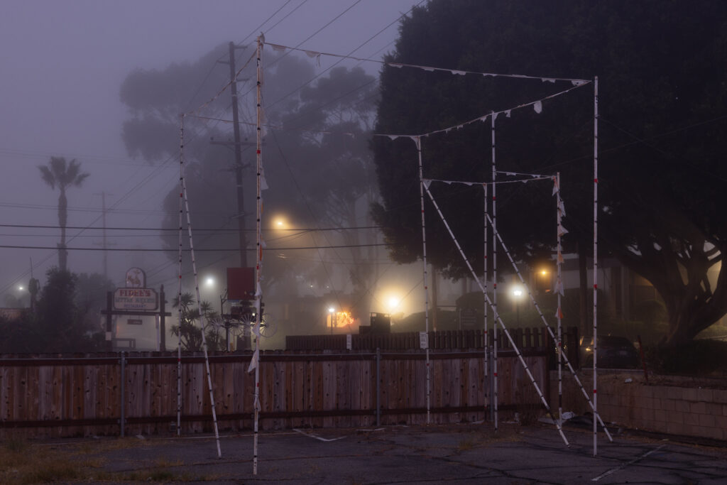 A dusk photograph of flags strung between white poles, illustrating the shape of a proposed building. In the background are Mexican restaurant signs.