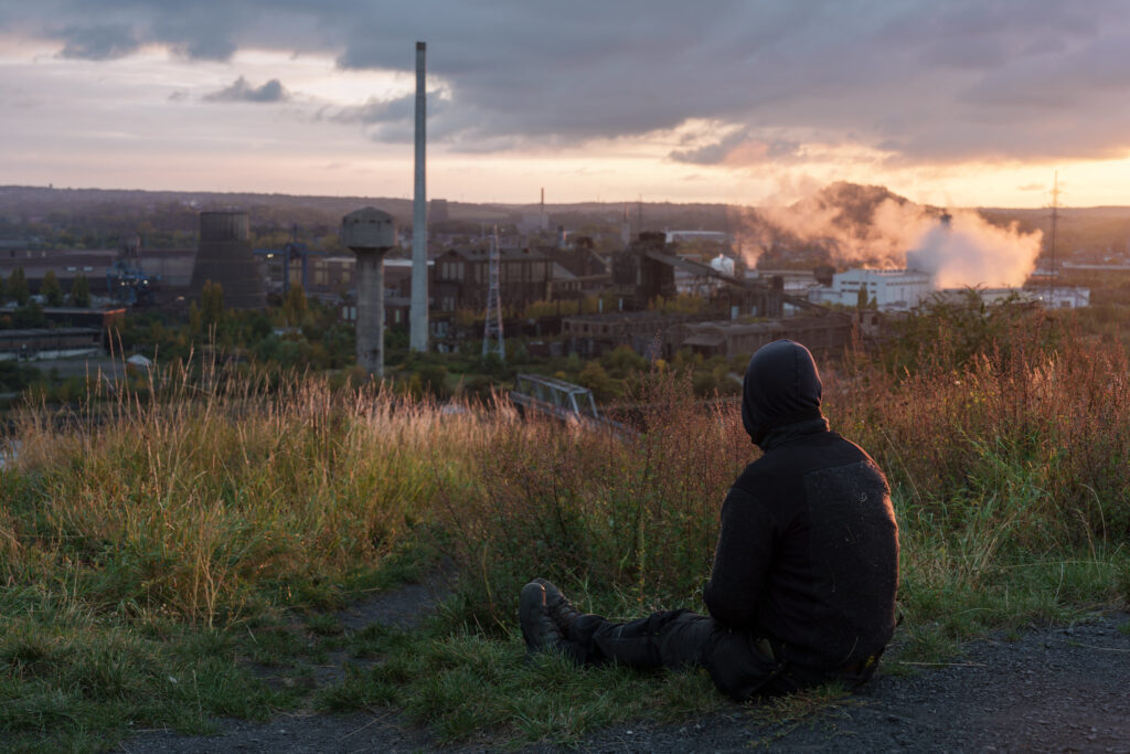 A photograph of a man sitting on a spoil tip overlooking the industrial Sambre River valley at sunset.