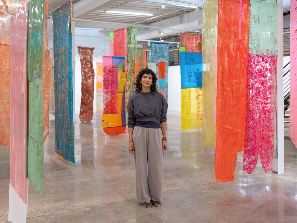 Artist Ivelisse Jimenez standing in her "Campo de Resonancia" installation of colorful hanging artwork.